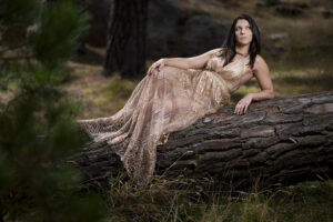 A woman reclining on a fallen tree trunk in the forest wearing a shimmery, golden dress.